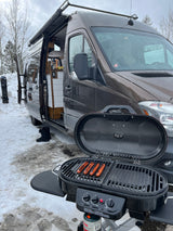 An RV rental van parked in snow, with a grill cooking hotdogs outside. Perfect for winter camping trips.