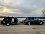 RV rental: Subaru Outback towing a black teardrop camper by a lake. Perfect for serene outdoor travel.