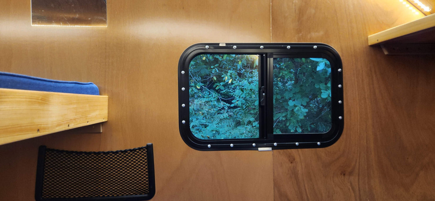 RV rental interior: bed with blue cushion, window showing trees, wood walls, net storage, and lights.