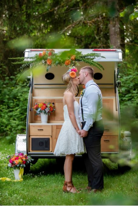 A joyful couple kisses beside a floral-decorated RV rental, a unique backdrop for their special outdoor event.