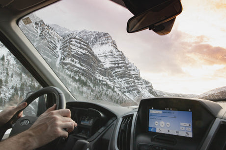 Driving an RV rental through majestic snowy mountains at sunset, hands on the wheel. Dashboard shows 59 mph.