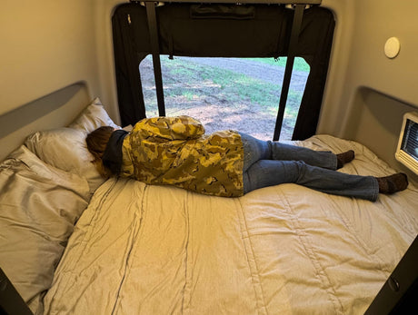 A person in camo and jeans relaxes face down on a bed in an RV rental, looking out the window.