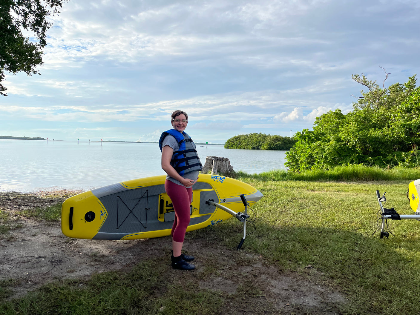 Smiling pregnant woman in a life vest by a yellow Hobie SUP near calm water and green banks. Enjoy nature with an RV rental!