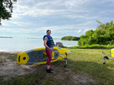Smiling pregnant woman in a life vest by a yellow Hobie SUP near calm water and green banks. Enjoy nature with an RV rental!