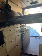Cozy rv rental interior: wooden storage, elevated bed with patterned blanket, fan, and a snowy view out back.