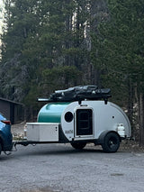 Compact teardrop RV rental attached to a blue SUV, parked on pavement in a forest setting. Equipped for outdoor camping.