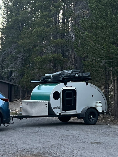 Compact teardrop RV rental attached to a blue SUV, parked on pavement in a forest setting. Equipped for outdoor camping.