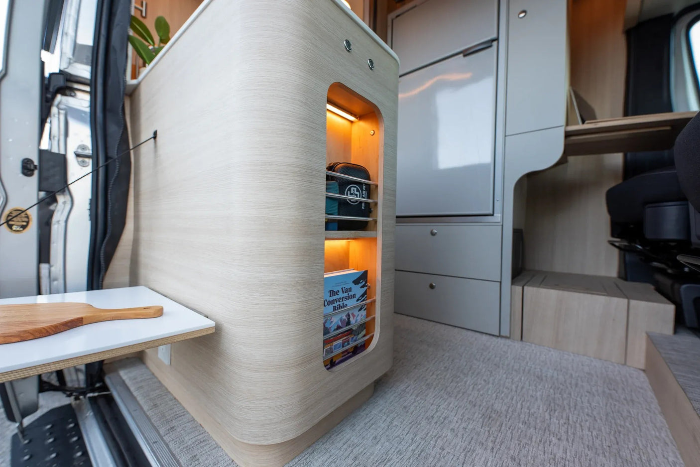 Stylish rv rental interior showcasing a light wood cabinet with illuminated storage for books & essentials, beside a fridge & work area.