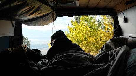 A person relaxes in an rv rental, looking out at vibrant fall foliage and distant mountains from a cozy bed, perfect for van life.