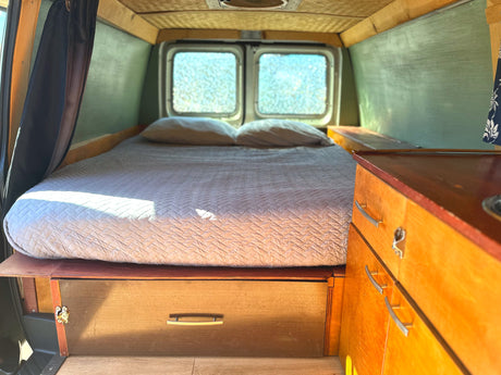 Cozy RV rental interior featuring a quilted bed, bright rear windows, and polished wooden cabinetry.