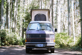 Front view of a Westfalia camper van, perfect for rv rental, parked in a sunny forest with its pop-top extended.