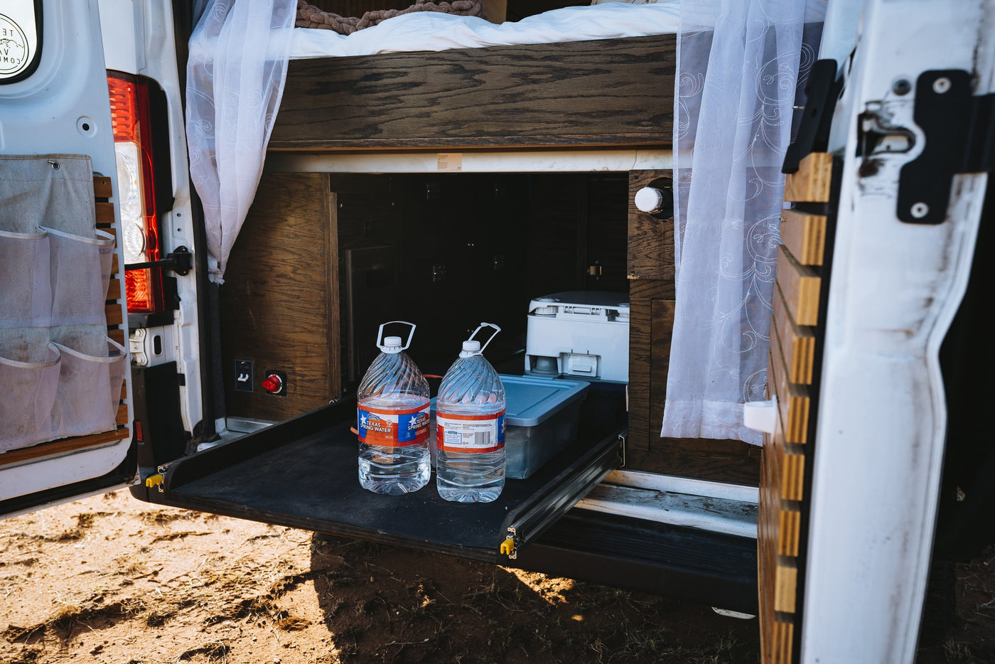 RV rental: rear of camper van, bed above storage. Pull-out tray holds two water jugs & a portable toilet. Sheer curtains visible. — photo 9