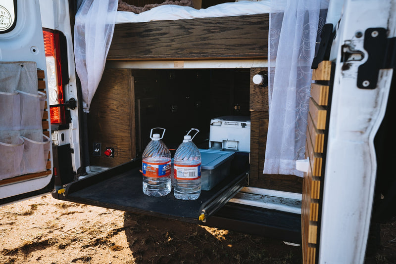 RV rental: rear of camper van, bed above storage. Pull-out tray holds two water jugs & a portable toilet. Sheer curtains visible. — photo 9
