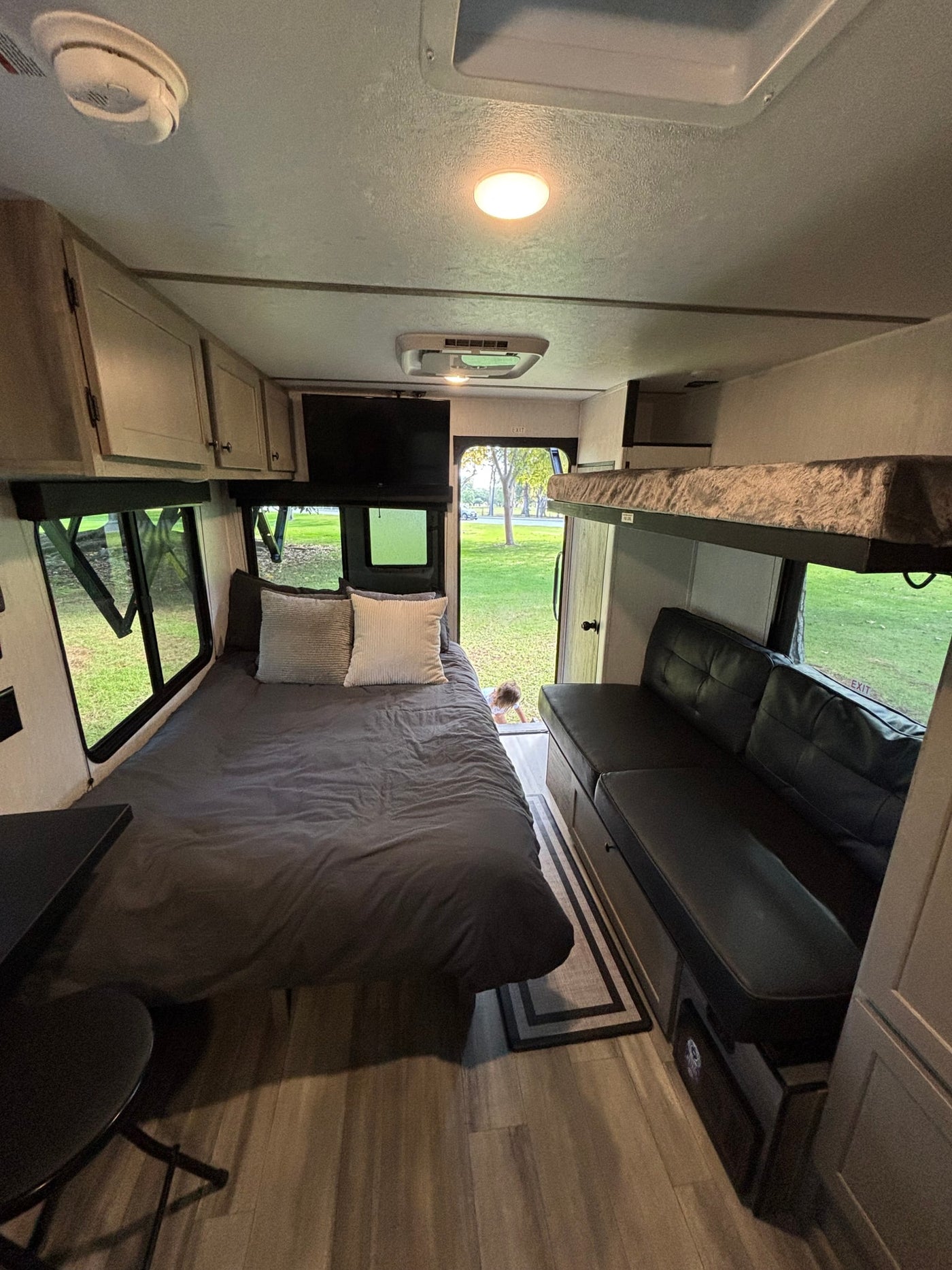 Cozy RV rental interior with bed, bunk beds, sofa, and TV. Features windows overlooking green outdoors and a child peeking. — photo 6