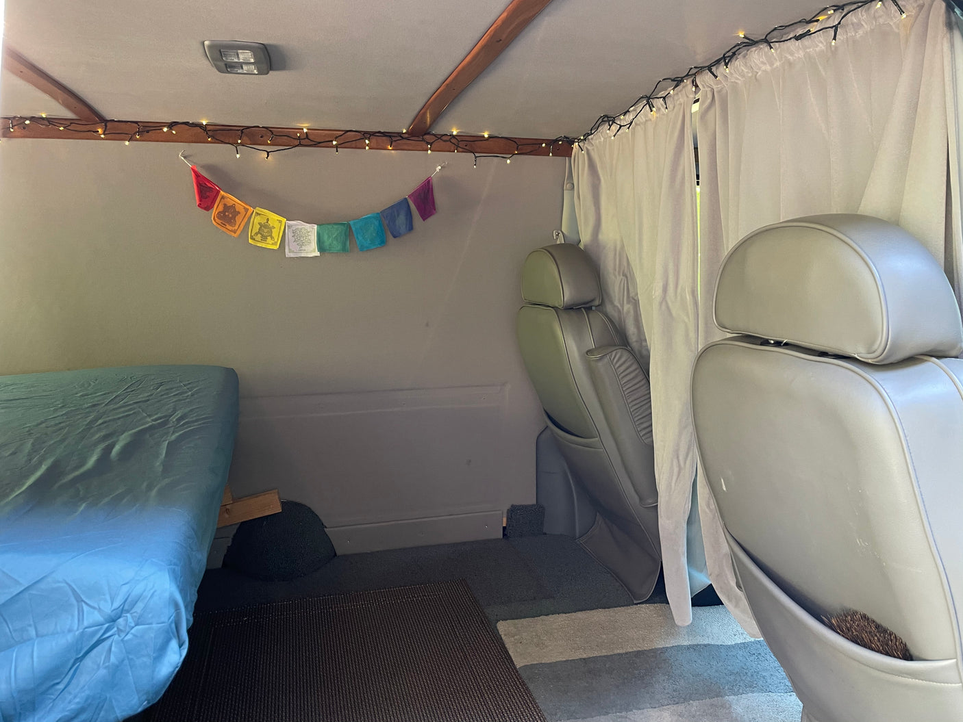 RV rental interior with bed, grey seats, white curtains, string lights, and prayer flags.