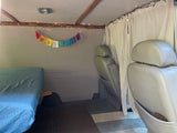 RV rental interior with bed, grey seats, white curtains, string lights, and prayer flags.