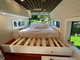 A custom RV rental interior showcasing a cozy bed with pull-out slats, white paneling, and an inviting forest backdrop.
