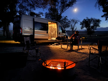 Cozy RV rental camping night with a person at a picnic table, by a campfire & camper van under moonlight.