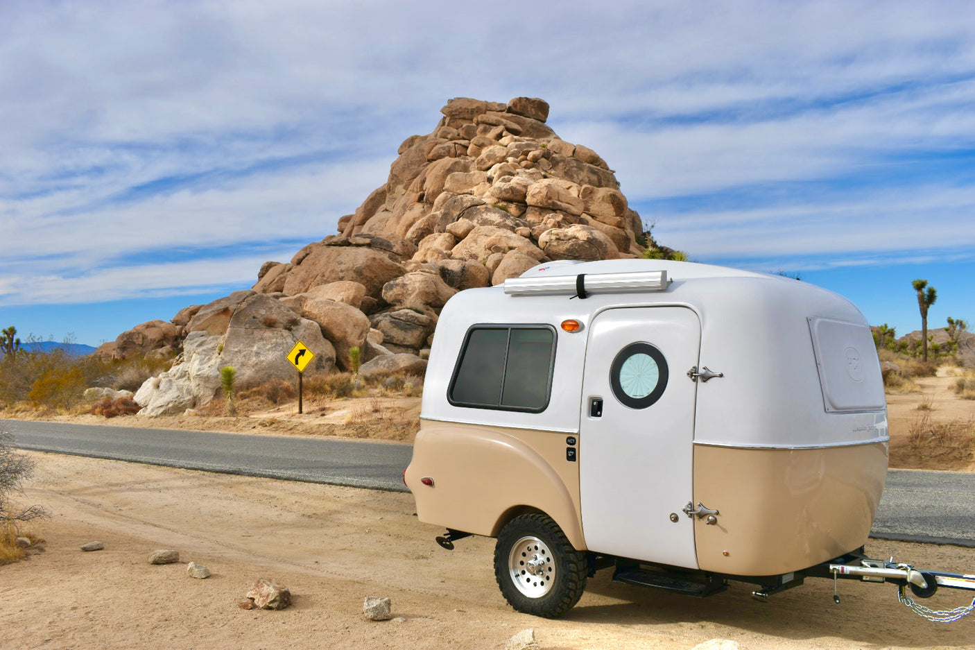 This two-tone compact RV rental, a teardrop trailer, is parked by a rocky desert road. Ideal for scenic travel & camping.