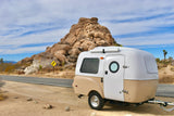 This two-tone compact RV rental, a teardrop trailer, is parked by a rocky desert road. Ideal for scenic travel & camping.
