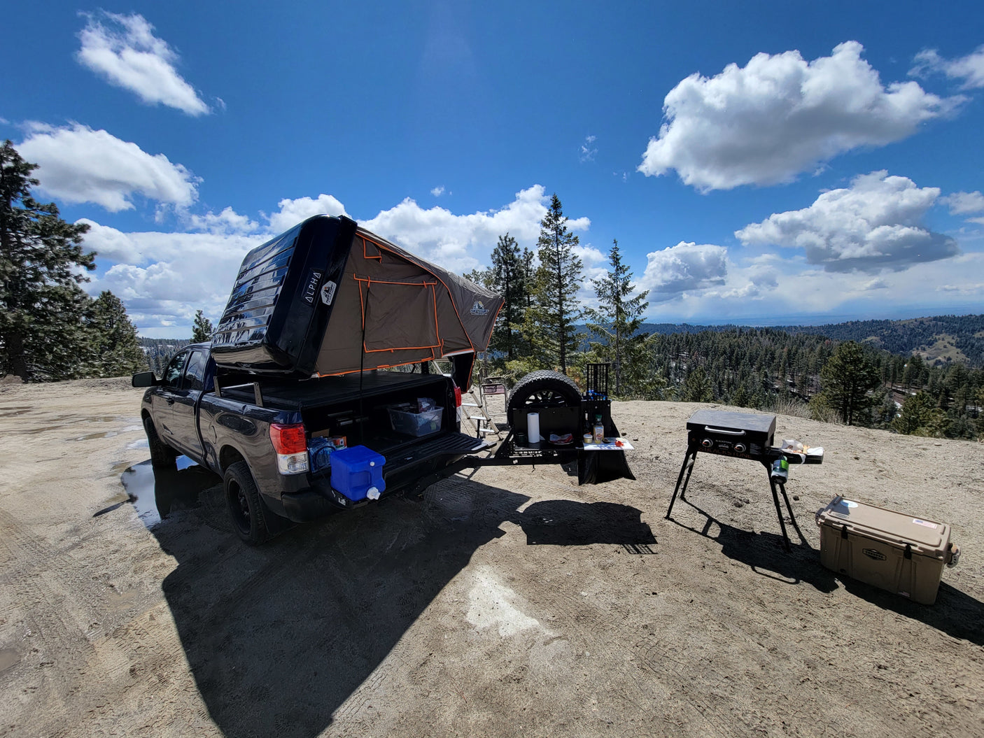 Truck with rooftop tent, grill, & cooler set up for camping in scenic mountains. An ideal RV rental alternative for adventure.