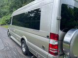 Silver Mercedes-Benz Sprinter Weekender RV for rental, showing its spacious side and rear with a spare tire carrier, parked by trees.