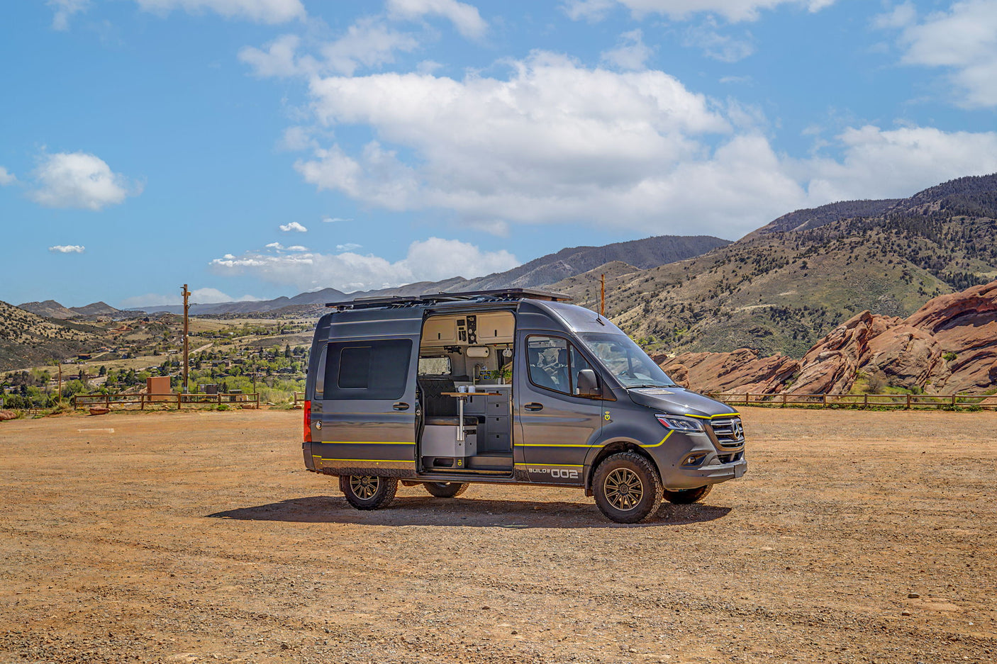 Adventure-ready grey Mercedes Sprinter rv rental camper van, side door open showing interior kitchen. Parked on dirt with mountains.