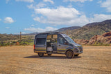Adventure-ready grey Mercedes Sprinter rv rental camper van, side door open showing interior kitchen. Parked on dirt with mountains.