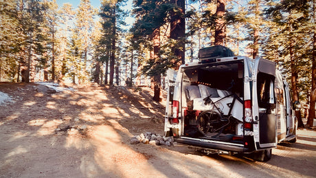 An RV rental sits in a sun-dappled forest, rear doors open revealing a cozy bed and bike inside for outdoor adventures.