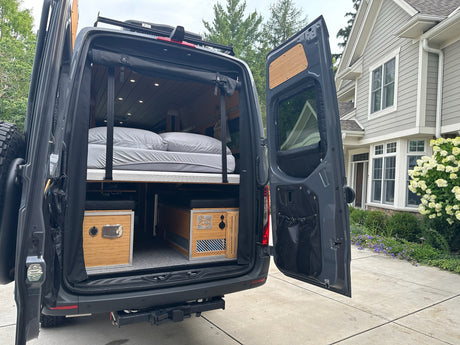 Rear view of an RV rental showing its interior: a raised bed with grey sheets and two bamboo storage units below.