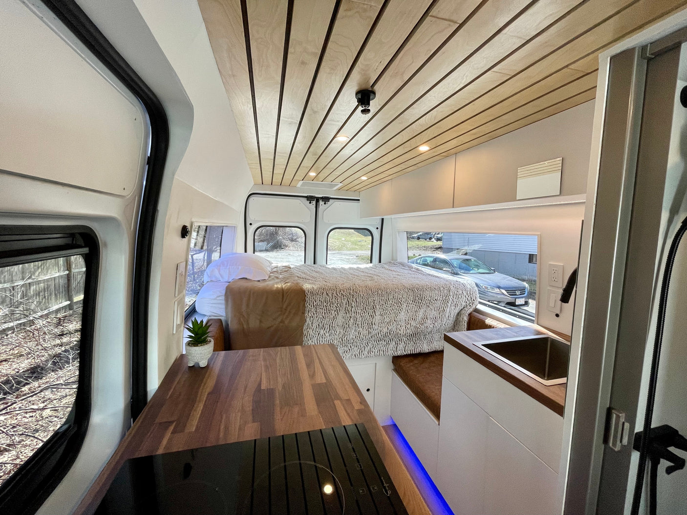 Modern RV rental interior showcasing a cozy bed, kitchenette with wood counters, and a stylish wooden slatted ceiling.