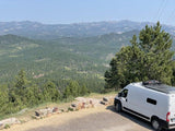 White rv rental van with solar panels overlooks a vast forested mountain landscape from a scenic viewpoint.