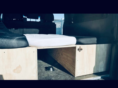 Inside an RV rental van: a cozy bed, wooden storage with a bottle opener, and a dog bowl below. Ready for adventures!