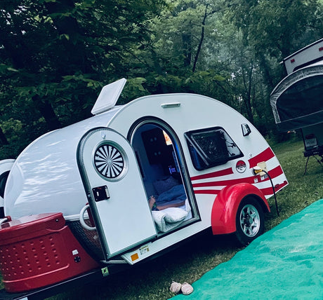 Cozy T@G teardrop RV rental with red stripes, parked in a wooded campsite, showcasing its compact, inviting interior.