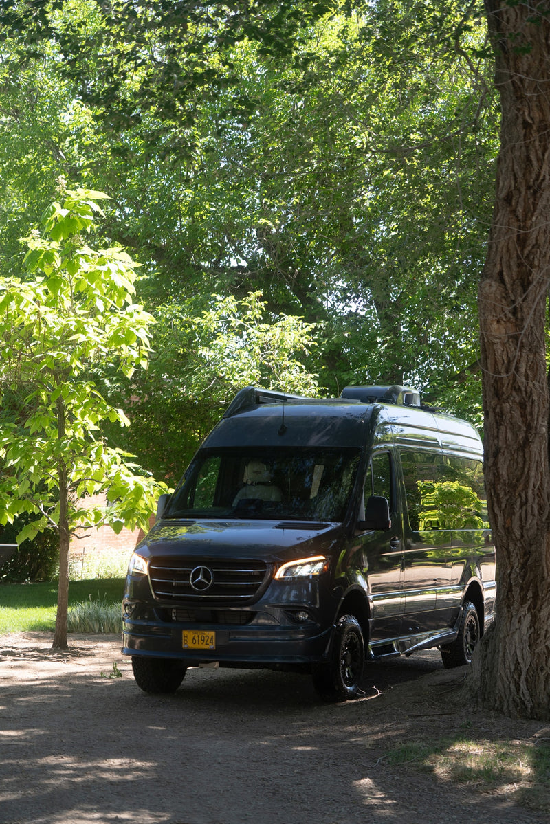 Dark gray Mercedes-Benz Sprinter rv rental van parked under dappled sunlight from lush green trees, ready for adventure. — photo 19