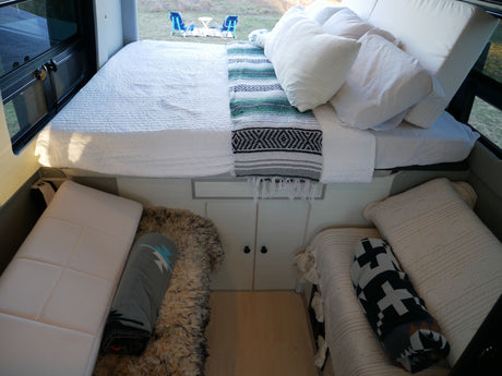 RV rental interior: A cozy bed overlooks outdoor chairs. Below are two side berths with blankets & pillows.