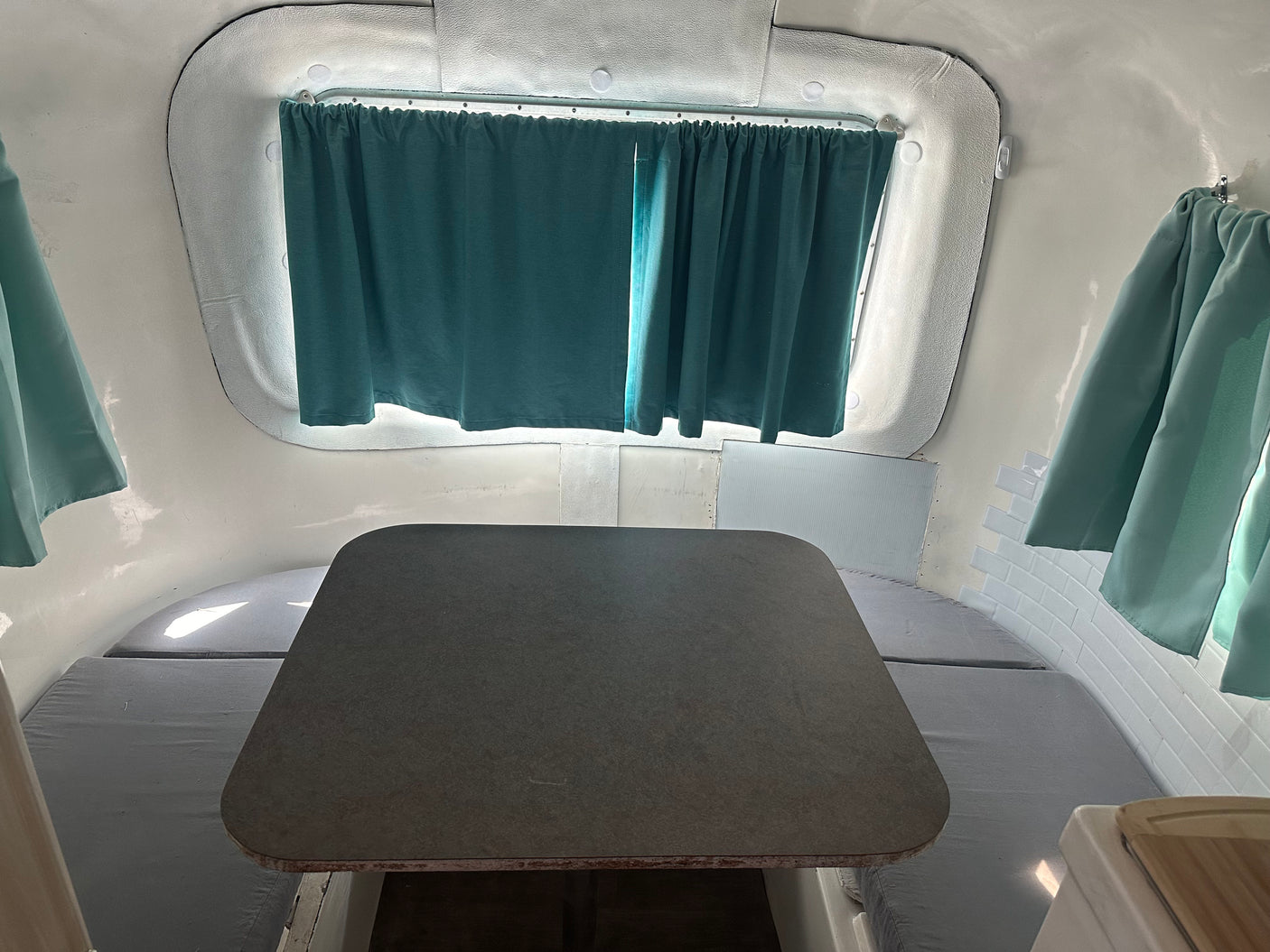 Compact RV rental interior featuring a gray table, benches with cushions, and teal curtains. Ideal for travel.