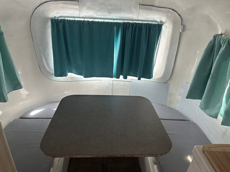 Compact RV rental interior featuring a gray table, benches with cushions, and teal curtains. Ideal for travel.
