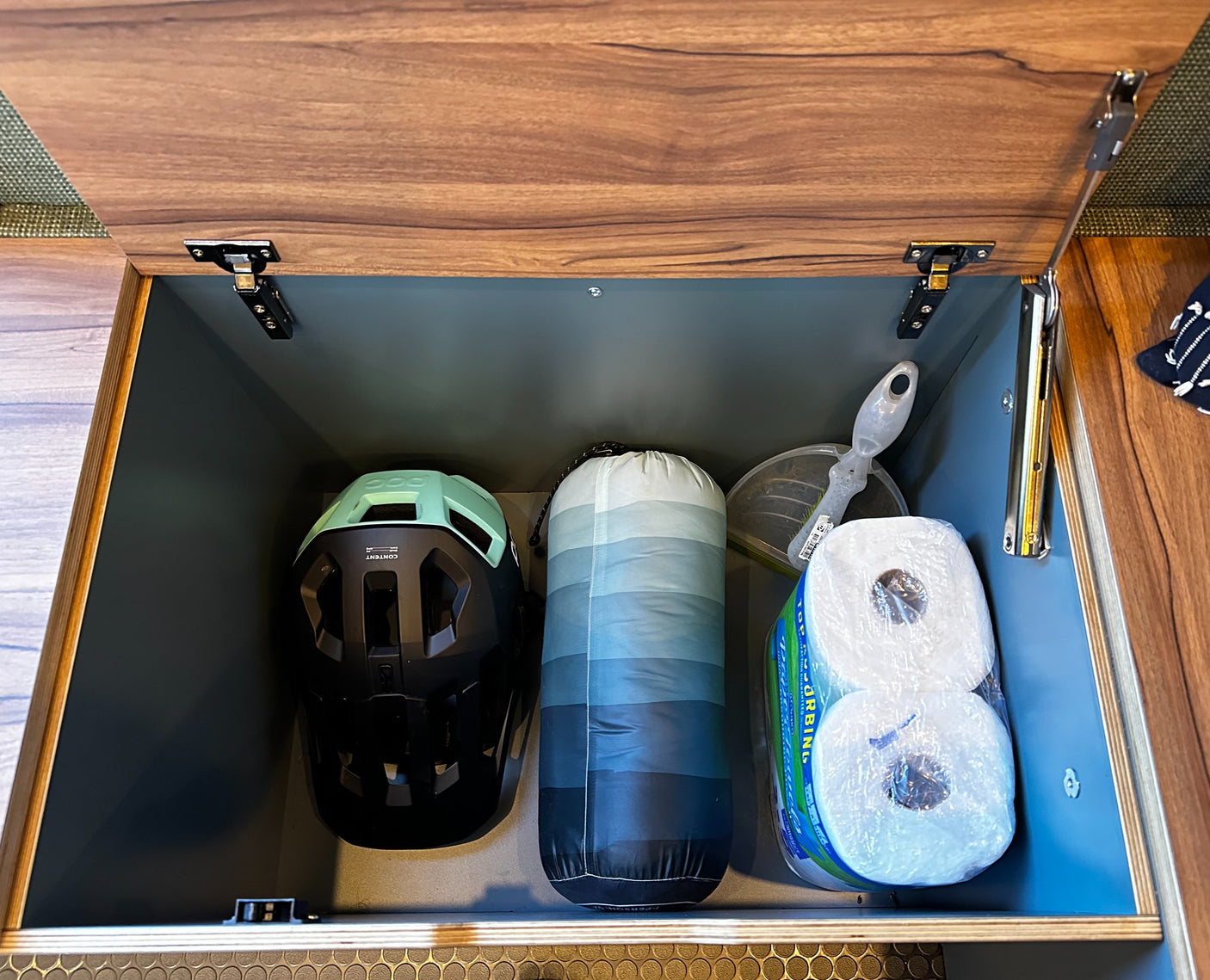 RV rental storage: Inside a dark grey RV compartment, a helmet, sleeping bag, & toilet paper are neatly stored.