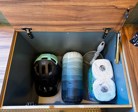 RV rental storage: Inside a dark grey RV compartment, a helmet, sleeping bag, & toilet paper are neatly stored.