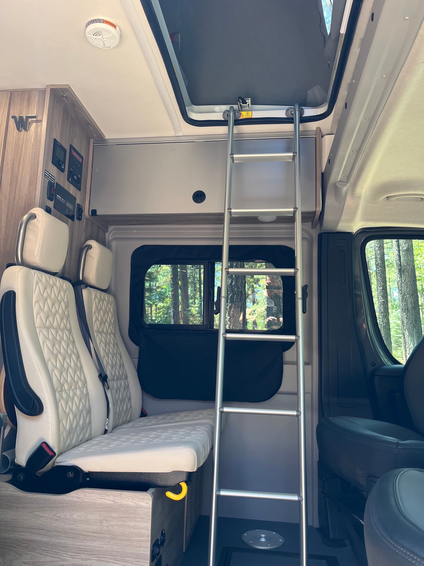 RV rental interior: Light-quilted passenger seats, control panel, ladder to upper bunk, and window view of trees.