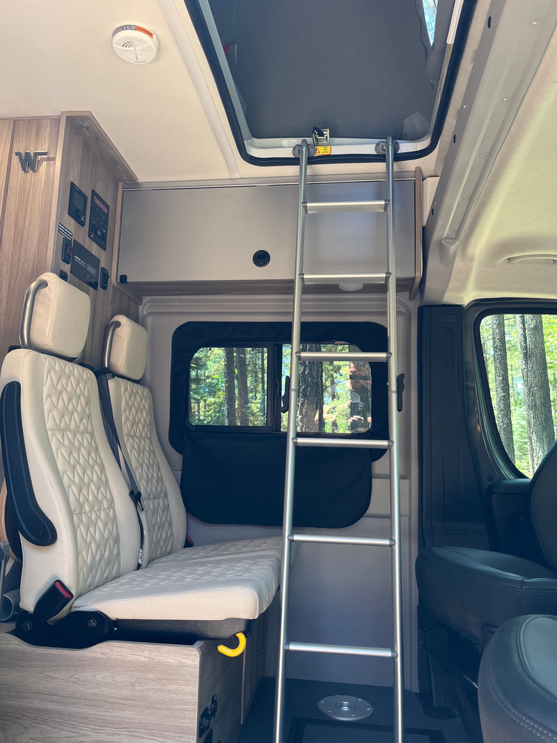 RV rental interior: Light-quilted passenger seats, control panel, ladder to upper bunk, and window view of trees. — photo 14