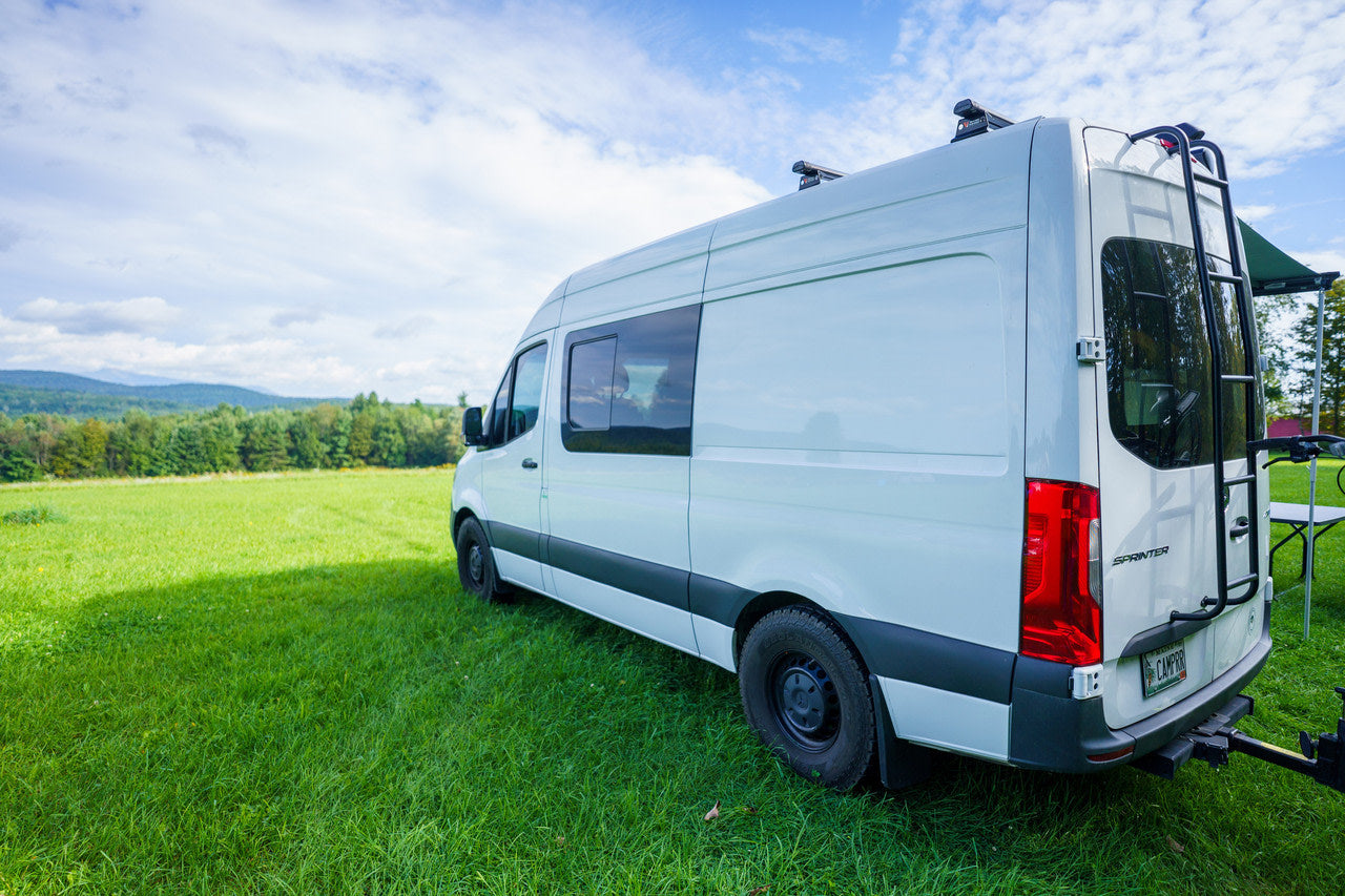 White Sprinter camper van, great for rv rental, parked in a green field with mountains under a cloudy sky.