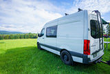 White Sprinter camper van, great for rv rental, parked in a green field with mountains under a cloudy sky.