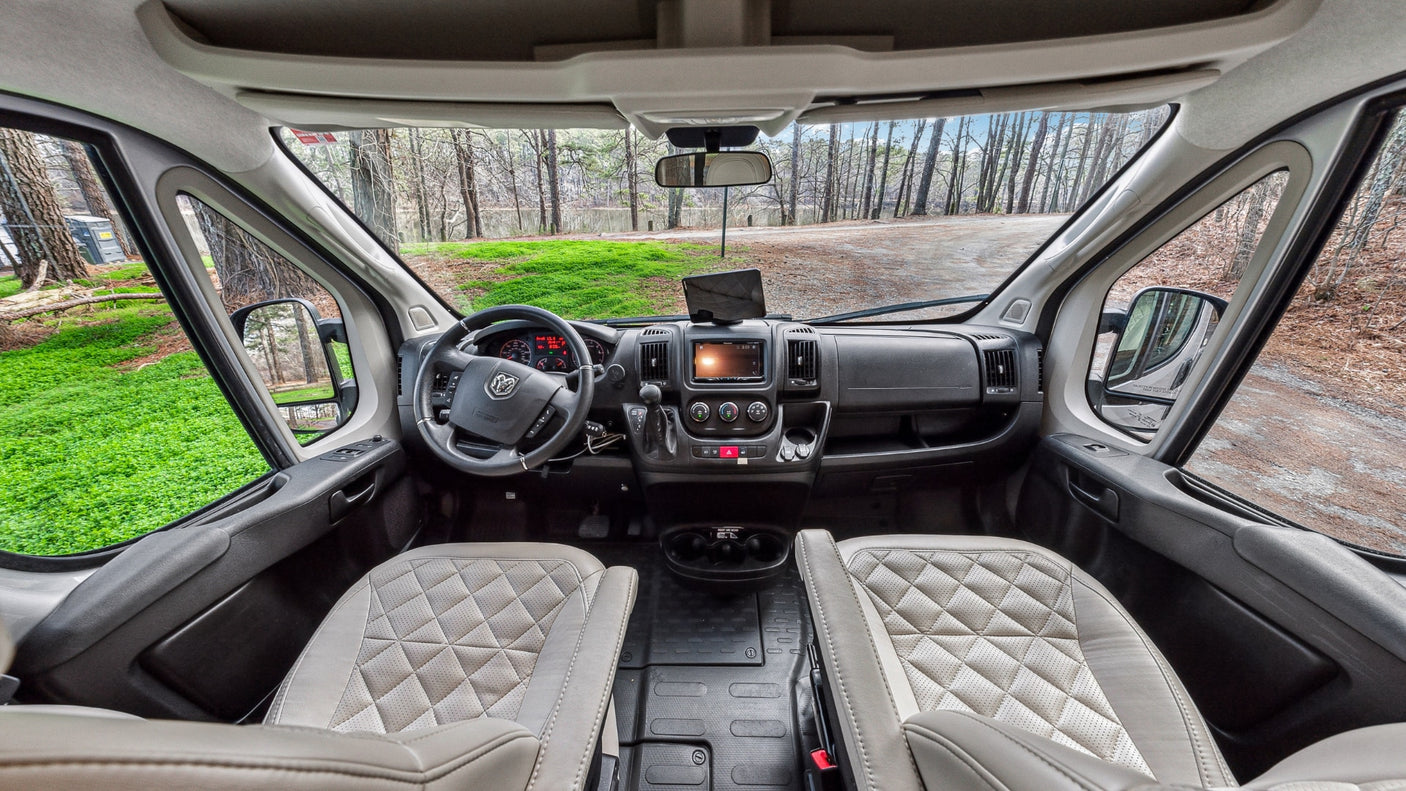 Spacious RV rental cockpit showing dashboard, steering wheel, quilted seats, and wooded view outside.