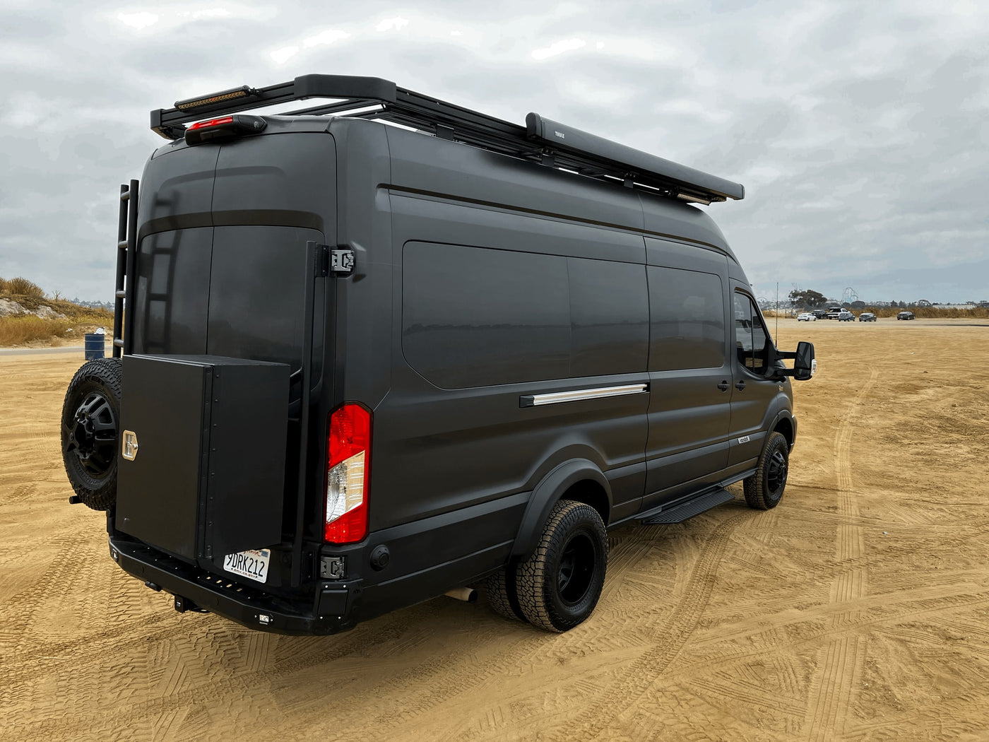 Black Ford Transit RV rental camper van on sand, with roof rack & awning. — photo 3
