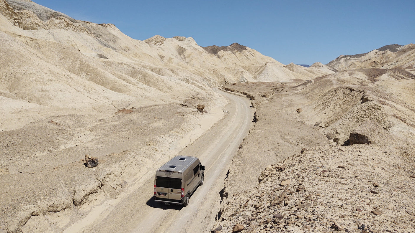 An RV rental navigates a winding dirt road through a vast, barren desert landscape of beige mountains under a clear blue sky.