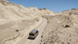 An RV rental navigates a winding dirt road through a vast, barren desert landscape of beige mountains under a clear blue sky.