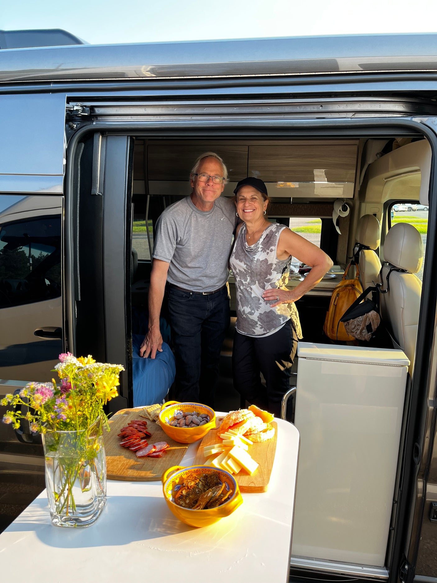 Smiling couple inside their RV rental van, enjoying an outdoor table set with snacks and flowers.