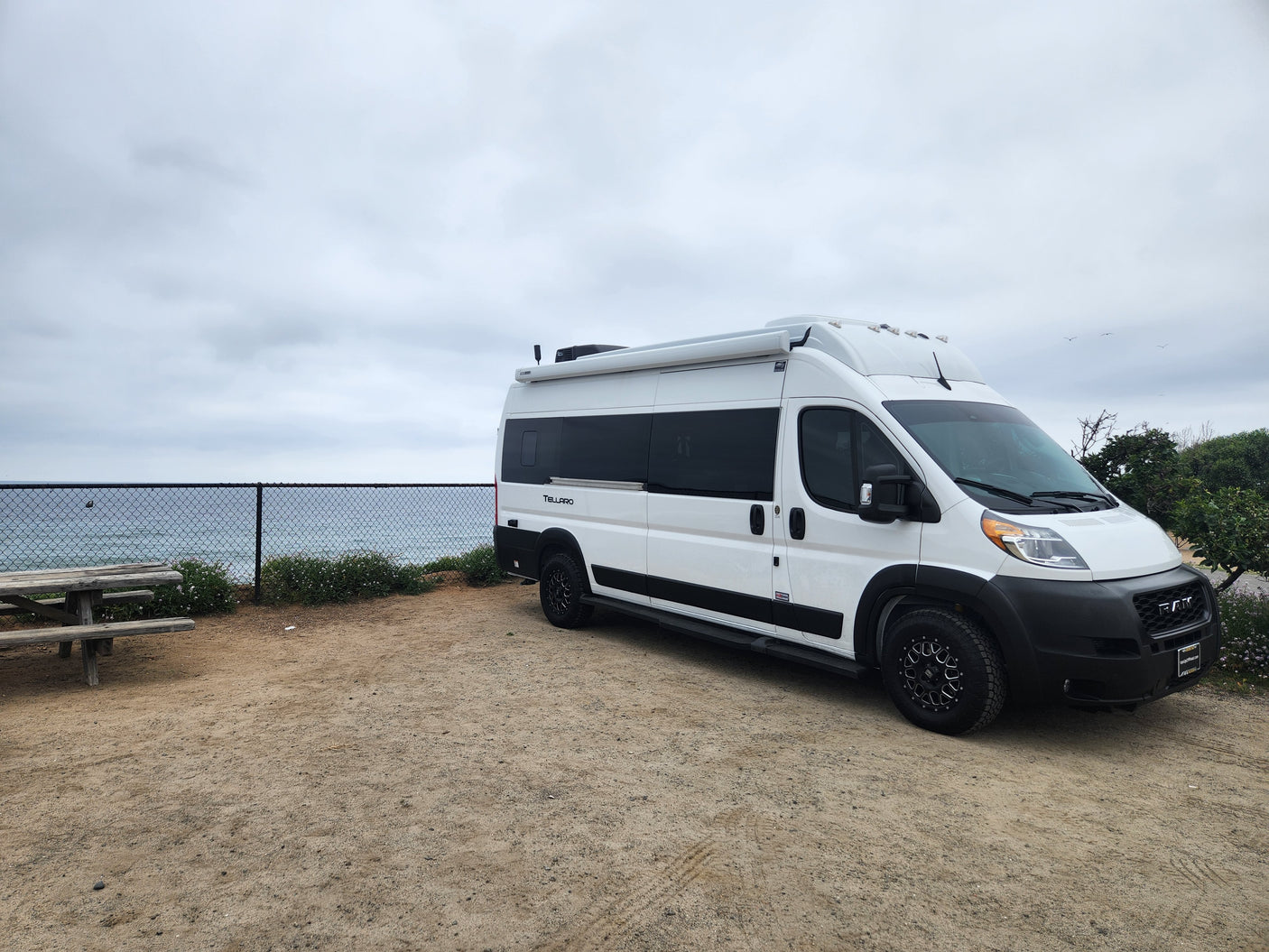 White **RV rental** van parked on a dirt lot overlooking the ocean, with a picnic table and cloudy sky.